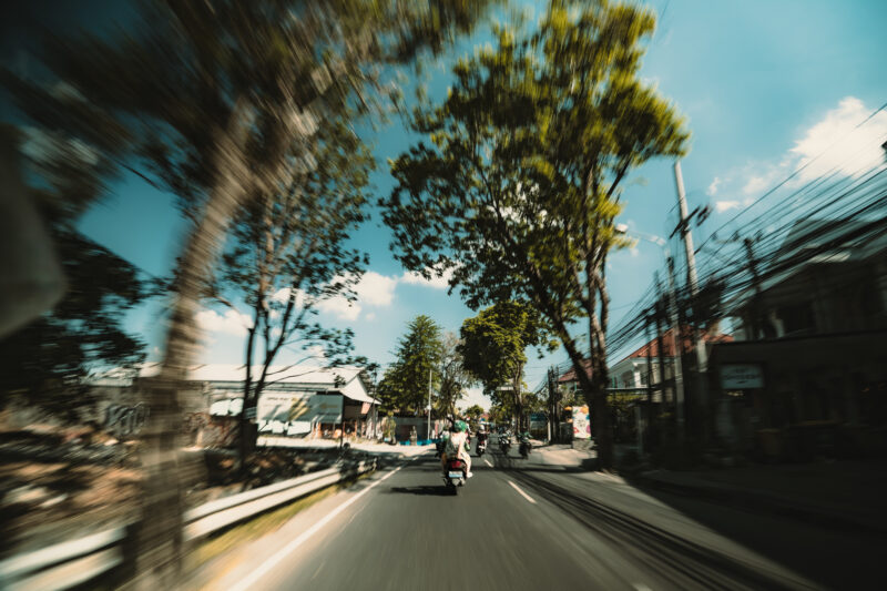 バリ島の市街地をバイクで走行する様子。中央にバイクに乗る二人乗りの姿があり、道路沿いには店舗や電線、住宅が並んでいる。